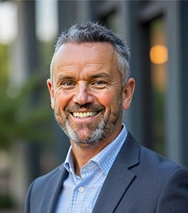 Head-and-shoulders portrait of a smiling man in a blue blazer outdoors.